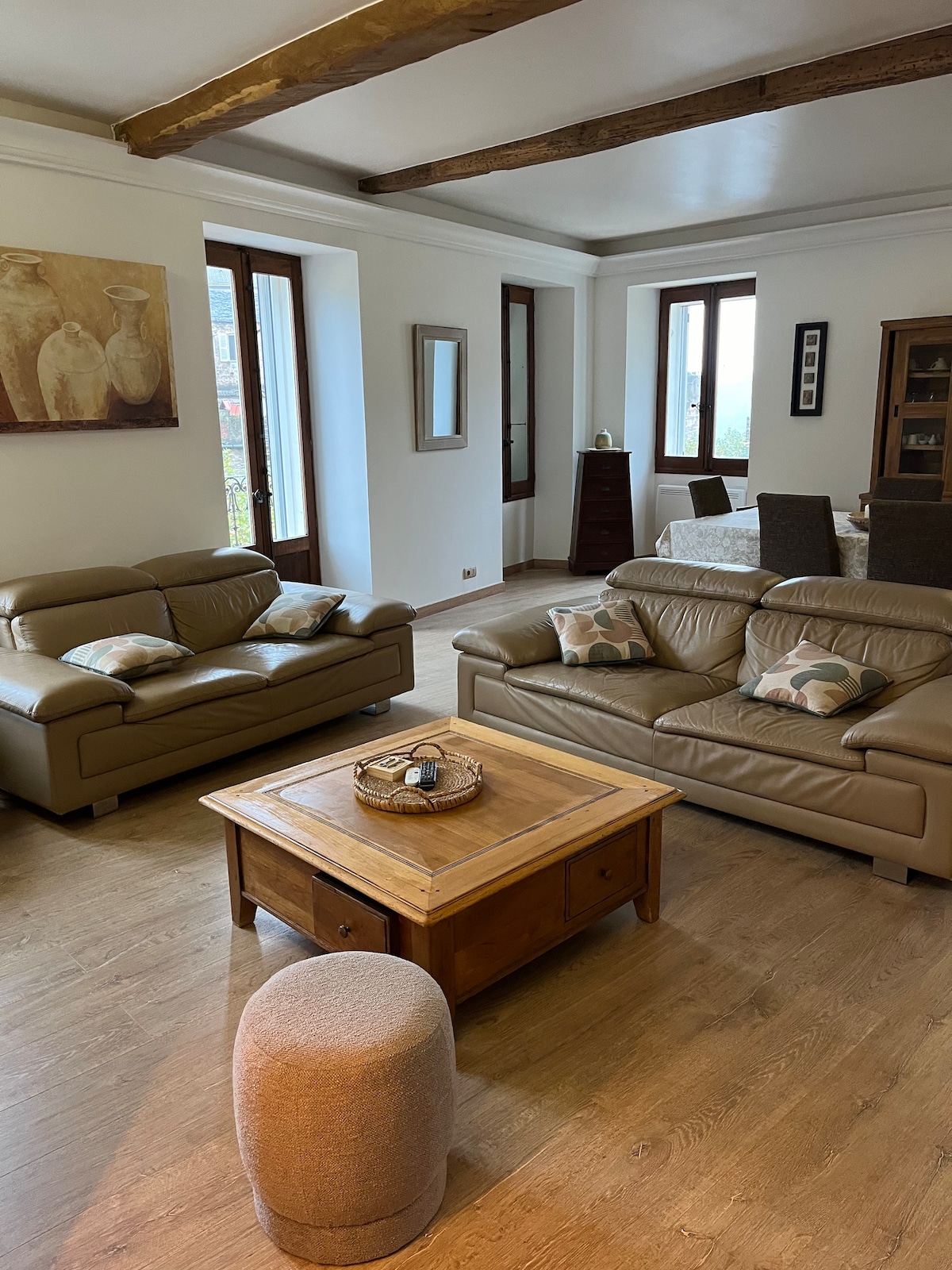 A spacious living area features two leather sofas, accented with decorative cushions. A wooden coffee table sits at the center, accompanied by a small ottoman. Large windows allow natural light to fill the room, and a dining table is visible in the background.