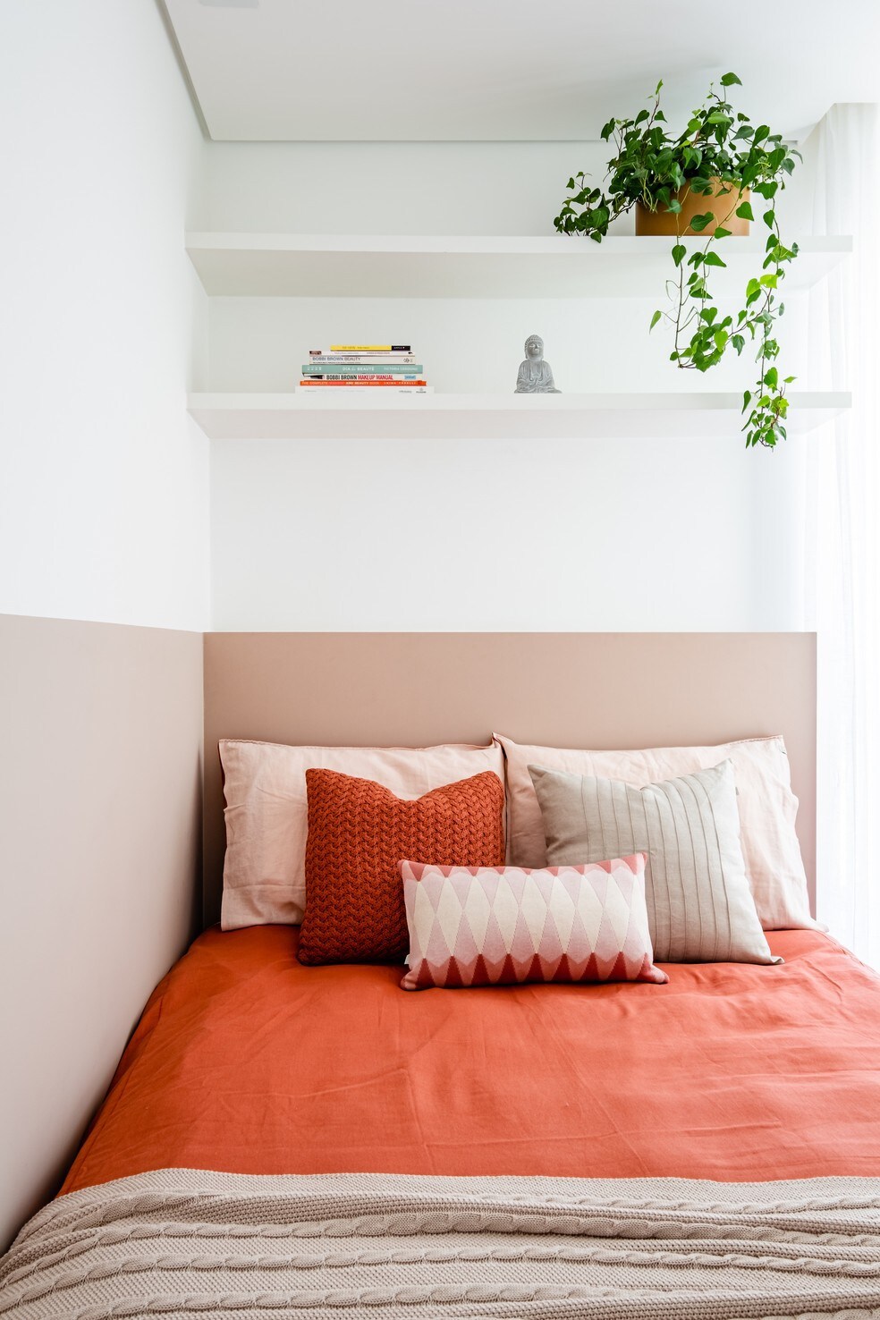A serene sleeping area is presented with a spacious bed dressed in a rust-colored coverlet. A variety of decorative pillows in soft hues and textures adorn the bed. Above, floating shelves display books and a small decorative statue, while a potted plant adds a touch of greenery.
