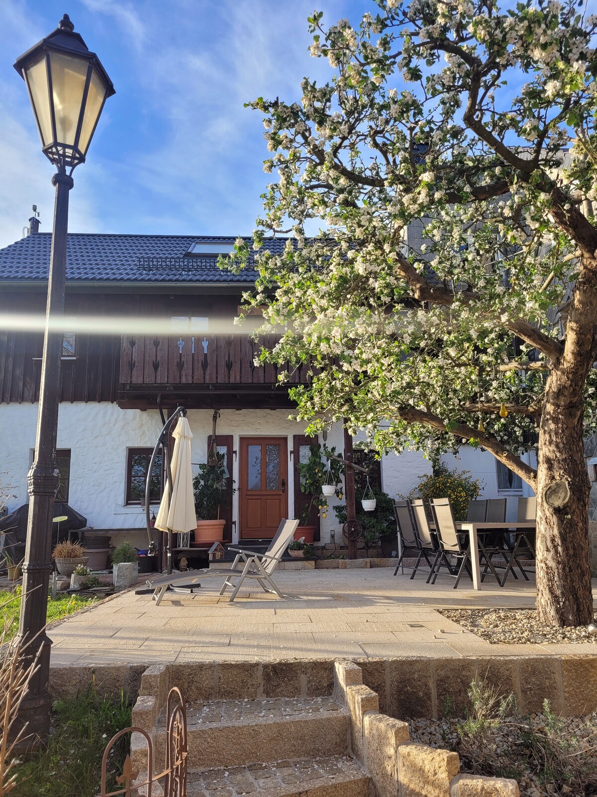 The exterior of the chalet features a well-maintained garden with a blooming tree and a patio area. A dining set is positioned for outdoor meals, while lounge chairs offer a space for relaxation. Soft light is cast from a lamp, creating a welcoming ambiance.