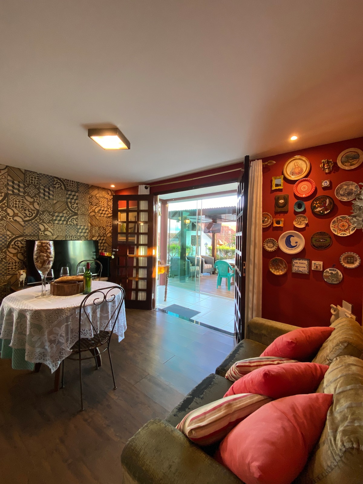 The inviting living area features a dining table set for two, surrounded by decorative plates on the red wall. A comfortable sofa adorned with cushions is positioned near the sliding glass doors, which open to a terrace area filled with greenery.