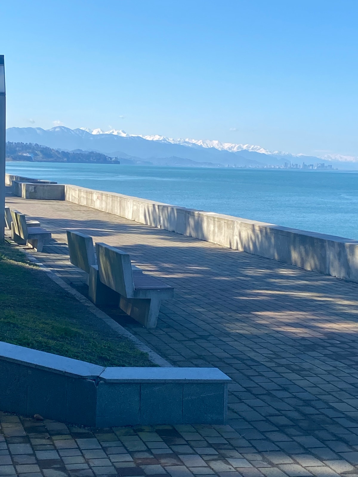 A scenic walkway is bordered by benches, offering views of the sea and distant mountains. Sunlight casts gentle shadows on the paved path, creating a serene environment for relaxation. The area appears inviting for leisurely strolls along the shore.