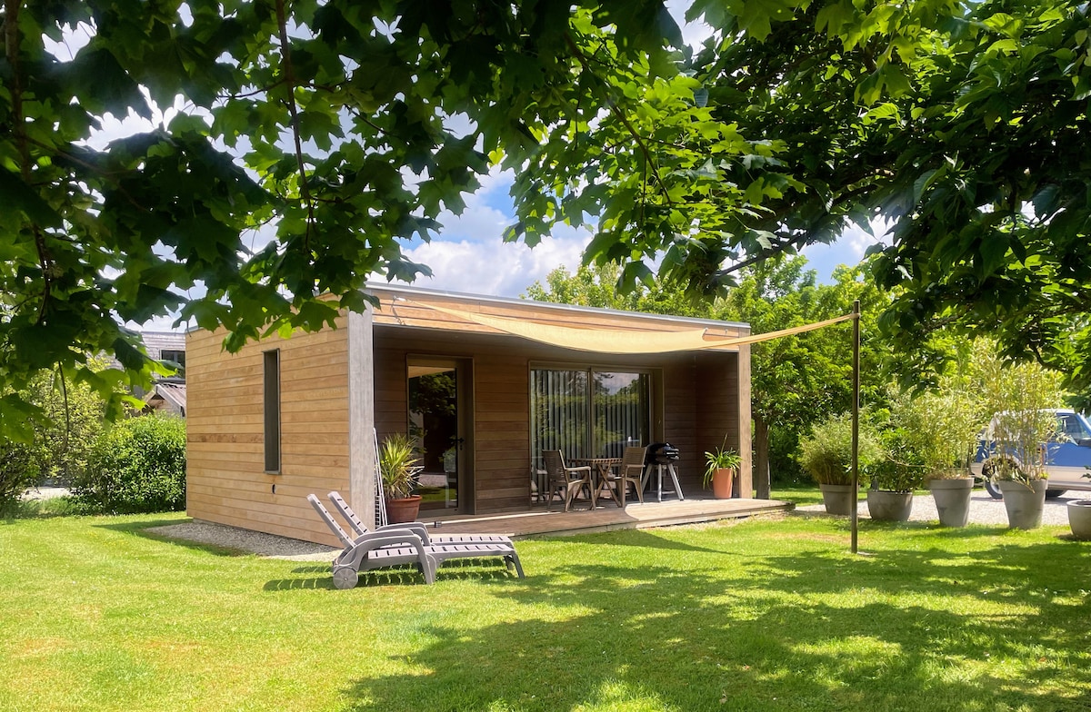 A modern wooden studio is surrounded by lush greenery and framed by trees. A small terrace features a table and chairs beneath a sunshade. Two lounge chairs are set on the well-kept lawn, providing a peaceful outdoor area for relaxation.