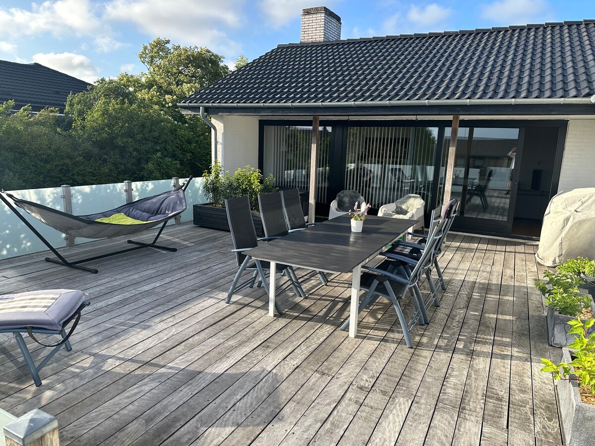 A spacious outdoor deck features a large dining table surrounded by several chairs. A hammock is positioned nearby, and various potted plants are arranged along the edges. Large sliding glass doors provide access to the interior, with a well-maintained wooden surface underfoot.