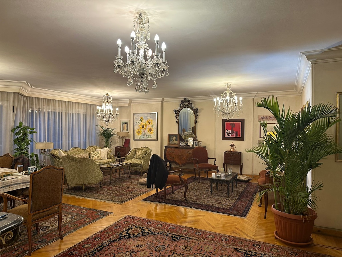 A spacious living area features elegant chandeliers casting soft light. Richly patterned rugs cover polished wood floors. A variety of seating options, including plush armchairs and a vintage sofa, are arranged around a decorative coffee table. Large windows are draped with sheer curtains, allowing natural light.