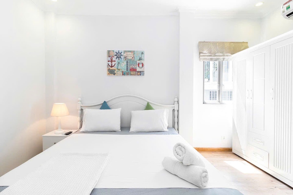 A bright studio room features a spacious bed with white linens and decorative pillows. Soft lighting is provided by a bedside lamp. A neatly arranged wardrobe is visible in the background, along with a framed artwork on the wall, enhancing the clean and inviting ambiance.