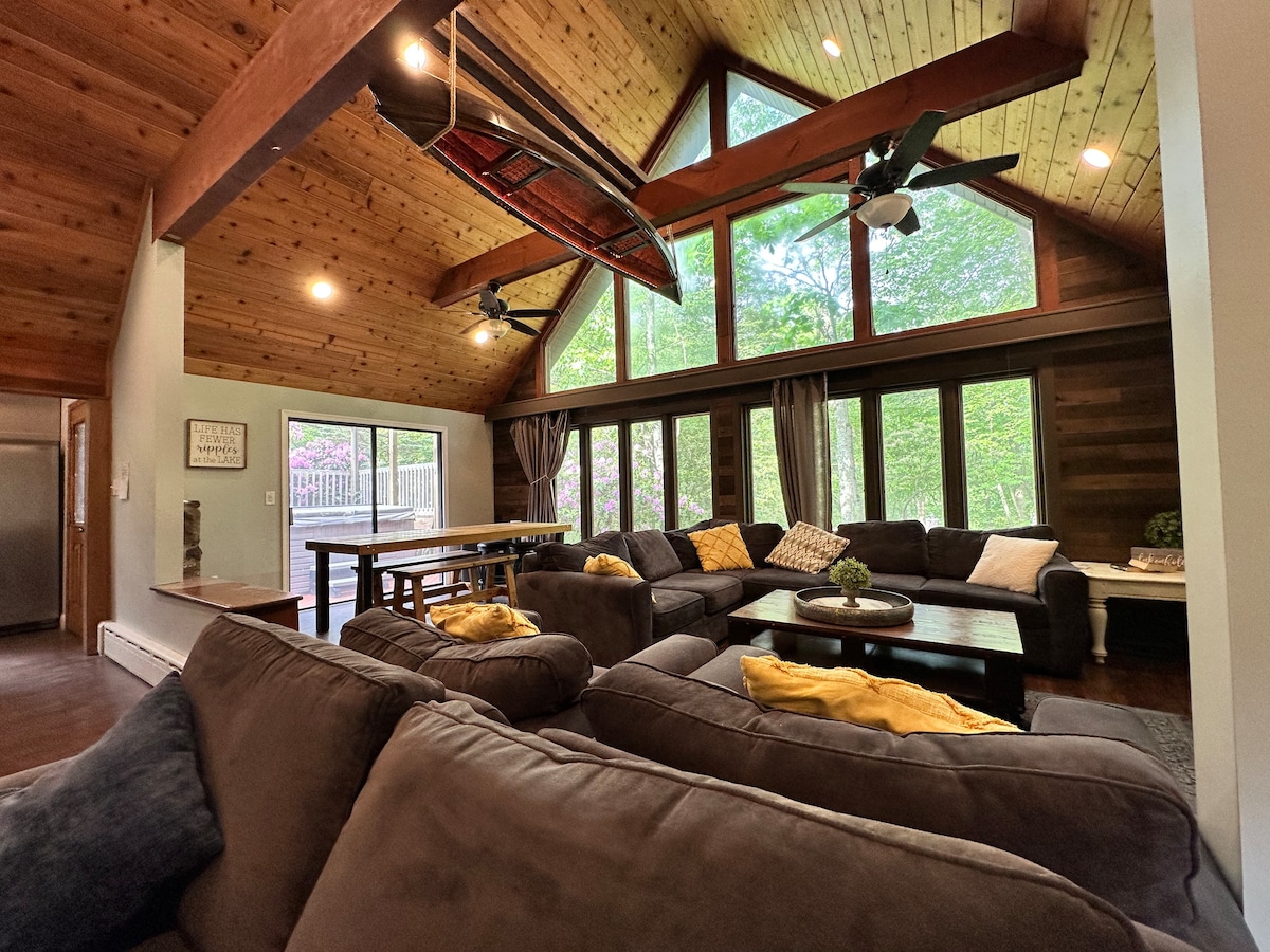 A spacious living area features vaulted ceilings with exposed wooden beams. Large windows invite natural light, offering views of the surrounding greenery. Comfortable seating is arranged around a central coffee table, complemented by decorative pillows. A dining table is positioned nearby, enhancing the open layout.