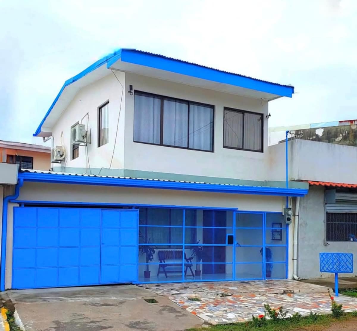 The exterior of a two-story house is displayed, featuring a blue roof and blue garage door. Large windows allow natural light into the upper level, while a stone-paved driveway leads to the entrance. Surrounding greenery enhances the welcoming appearance of the home.