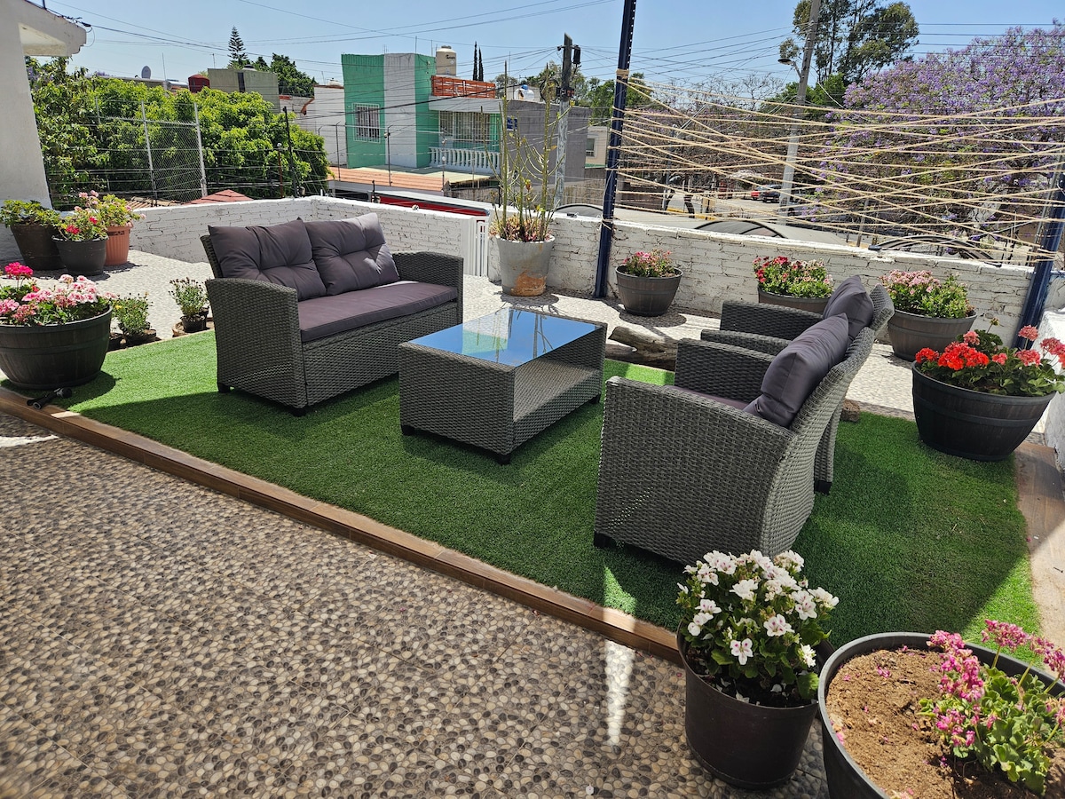 An outdoor terrace is presented, featuring comfortable seating arrangements with a coffee table at the center. The area is enhanced by lush artificial grass and surrounded by vibrant flower pots, providing a serene environment for relaxation.