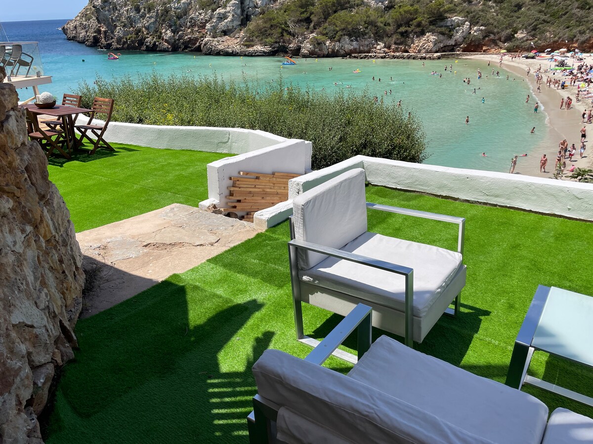 A terrace area is furnished with modern outdoor seating, featuring two white armchairs and a glass-topped table. The lush green carpet mimics grass, and a scenic view of the sandy beach with clear water and rocky cliffs is visible in the background.