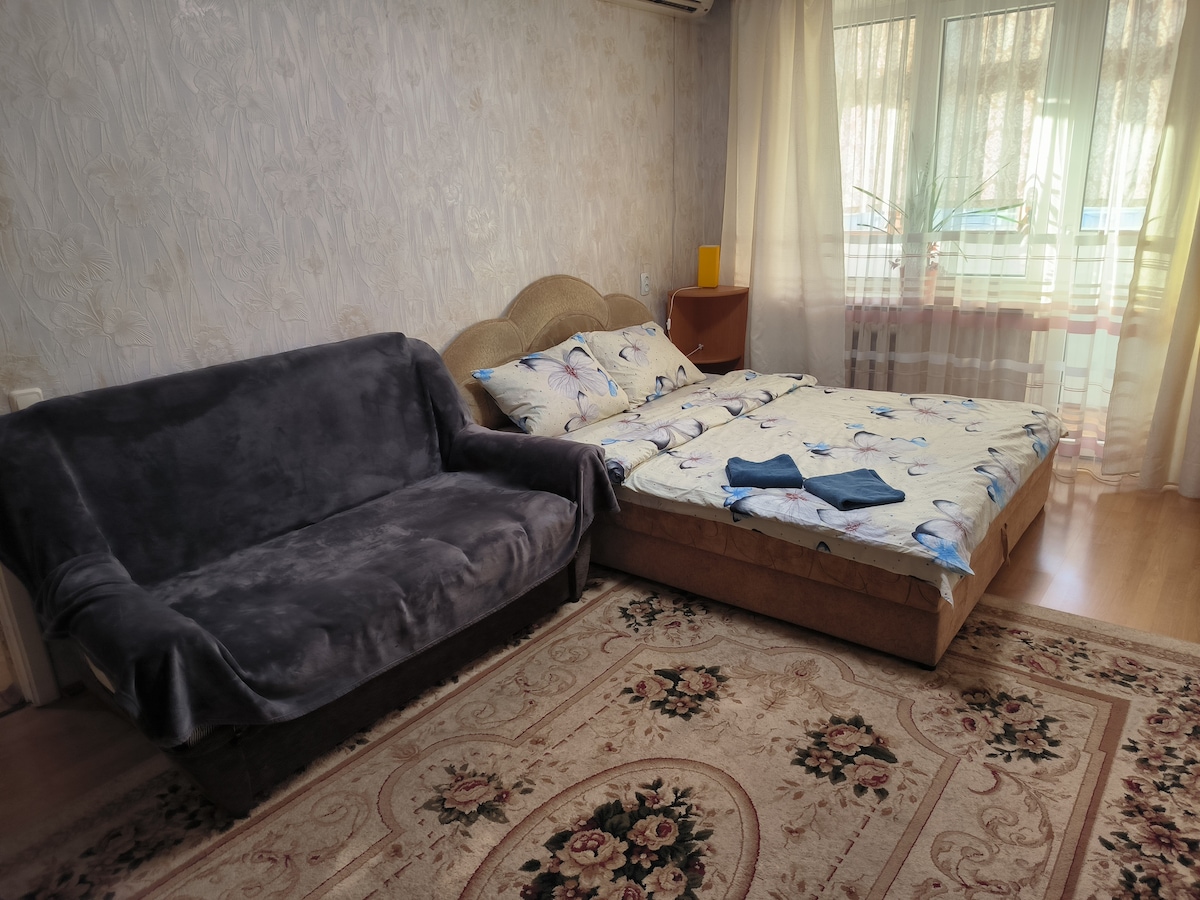 A comfortable sleeping area features a spacious bed with patterned bedding and a coordinated blue throw placed at the foot. A gray sofa is located adjacent to the bed. Cream curtains allow natural light to filter in, illuminating the light-toned walls and floral rug.