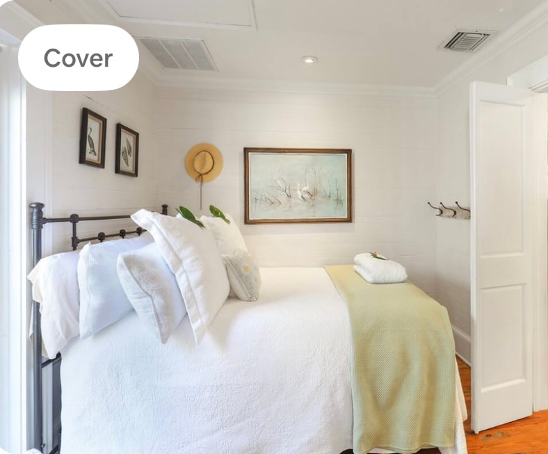 A cozy sleeping area features a full bed with crisp white linens and a soft pastel throw. Natural light highlights the room's bright decor, complemented by a framed artwork and a stylish straw hat mounted on the wall. A towel is neatly arranged at the foot of the bed.