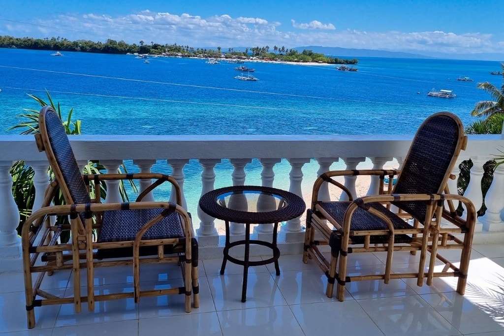 A serene balcony setting features two rattan chairs positioned alongside a round table, overlooking a clear blue ocean. The horizon showcases gentle waves and distant islands under a bright sky, creating a tranquil environment for relaxation.