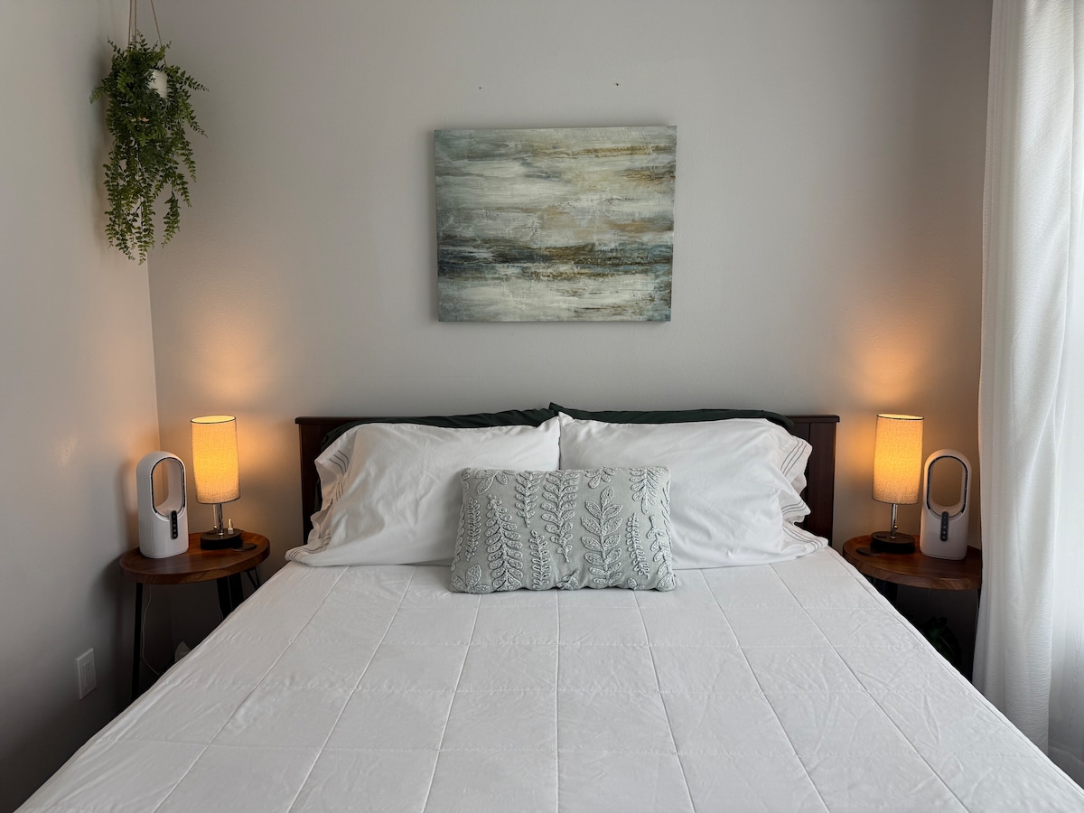 The sleeping area features a spacious bed with crisp white linens and a decorative pillow in the center. Soft lighting is provided by two bedside lamps on wooden tables, and a piece of abstract wall art adds a subtle touch above the bed. Greenery is present in the form of a hanging plant.