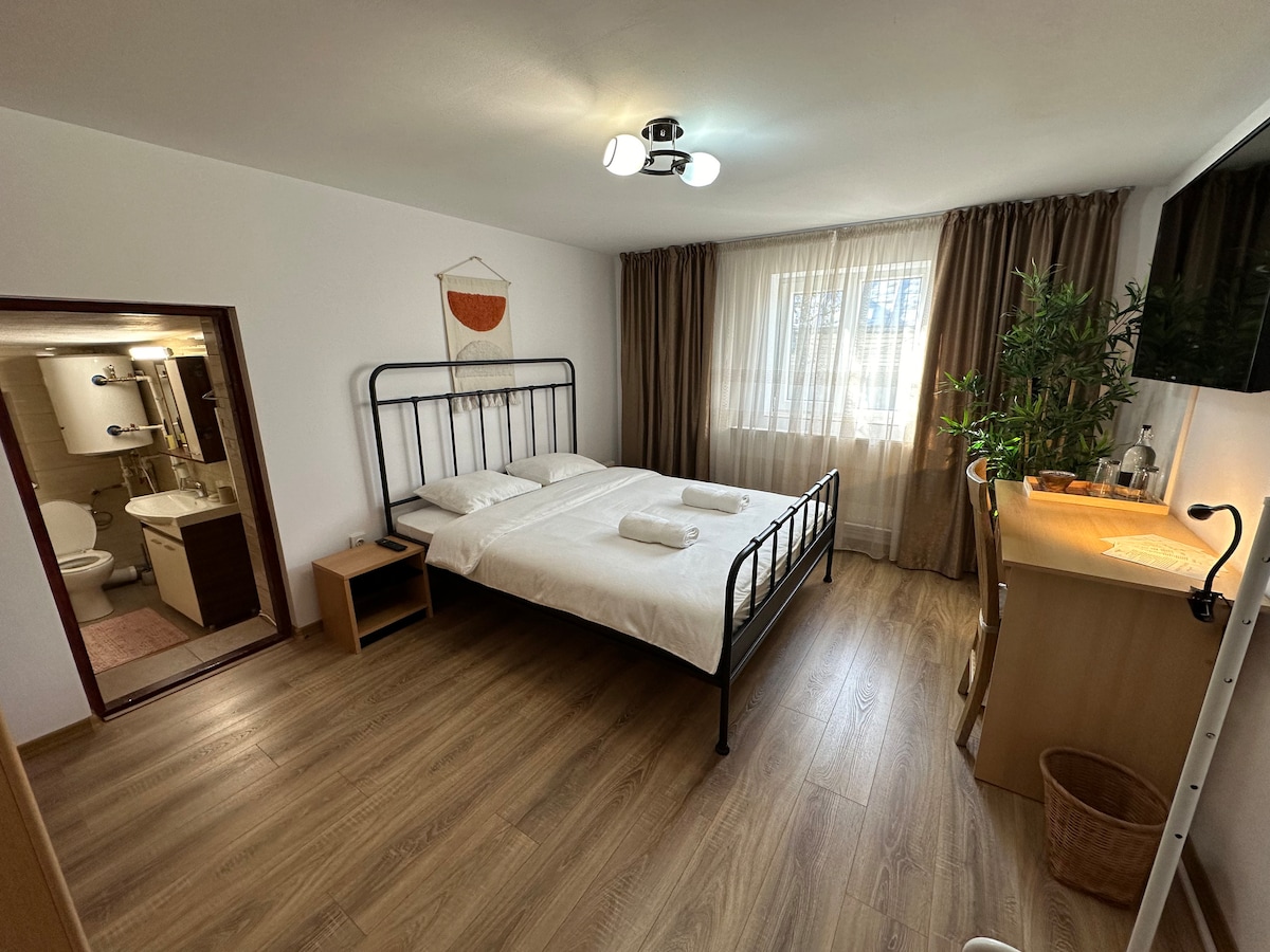 A bright and airy bedroom features a spacious bed with crisp linens and fluffy towels placed neatly. A small desk is positioned next to a window that allows natural light to fill the room. An entrance leads to an en-suite bathroom, enhancing convenience.