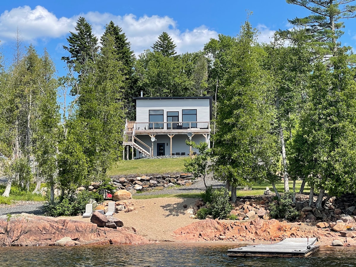 A lakeside retreat is surrounded by lush greenery and tall trees, with a sandy beach leading to the water. The two-story house features large windows on the upper level, allowing ample natural light, and a spacious deck overlooking the serene landscape.