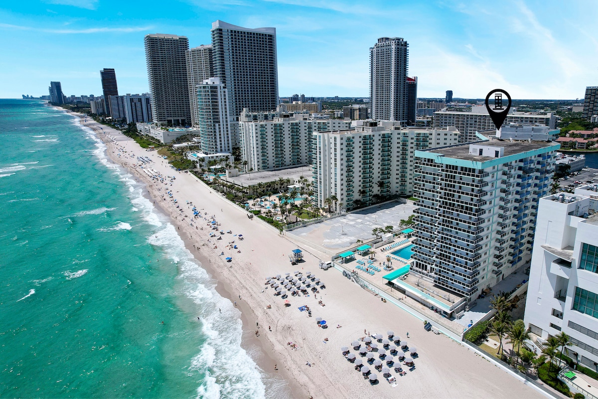 Aerial view capturing Hallandale Beach with sandy shoreline and vibrant turquoise waters. The oceanfront condo is depicted at the forefront, surrounded by several high-rise buildings, with beachgoers and umbrellas dotting the shore, offering a sense of the beach's lively atmosphere.