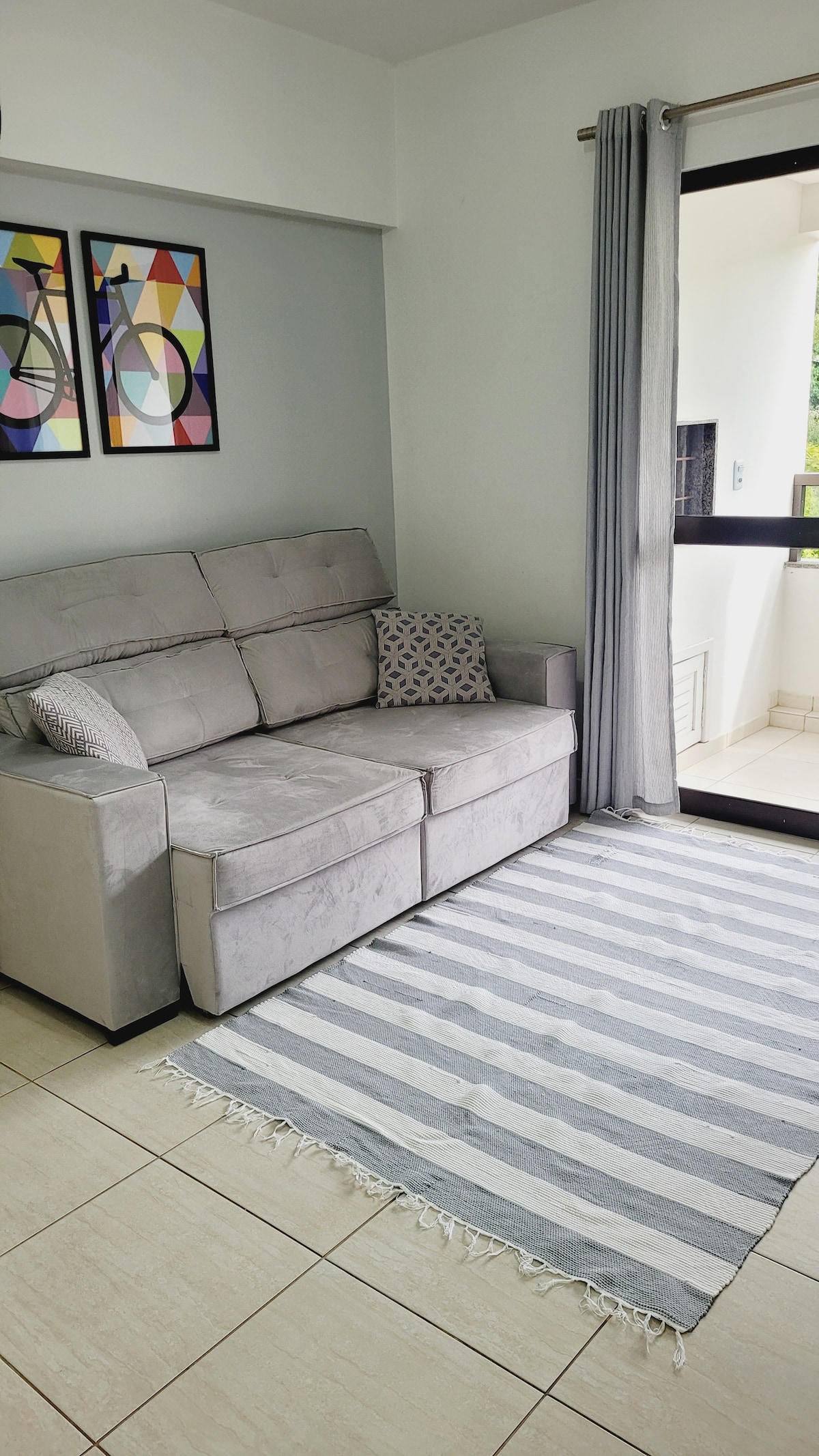 A light-colored sofa with decorative cushions is positioned against a wall, accompanied by two framed artworks featuring abstract designs. Natural light enters through a balcony door, which leads to an outdoor space. A striped area rug adds texture to the tiled floor.