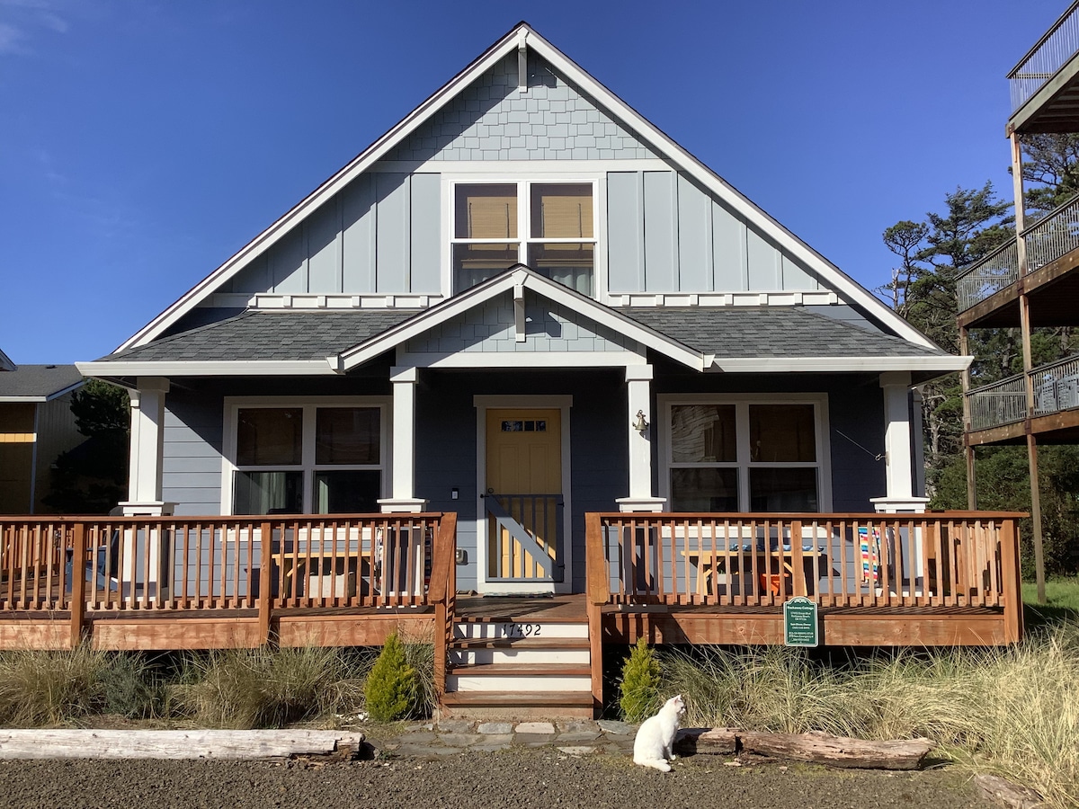 The exterior of Rockaway Cottage is presented, featuring a blue-grey facade and a welcoming yellow front door. A spacious wrap-around deck with wooden railings houses several seating areas. A small dog is seated near the entrance amidst landscaped grass and gravel.