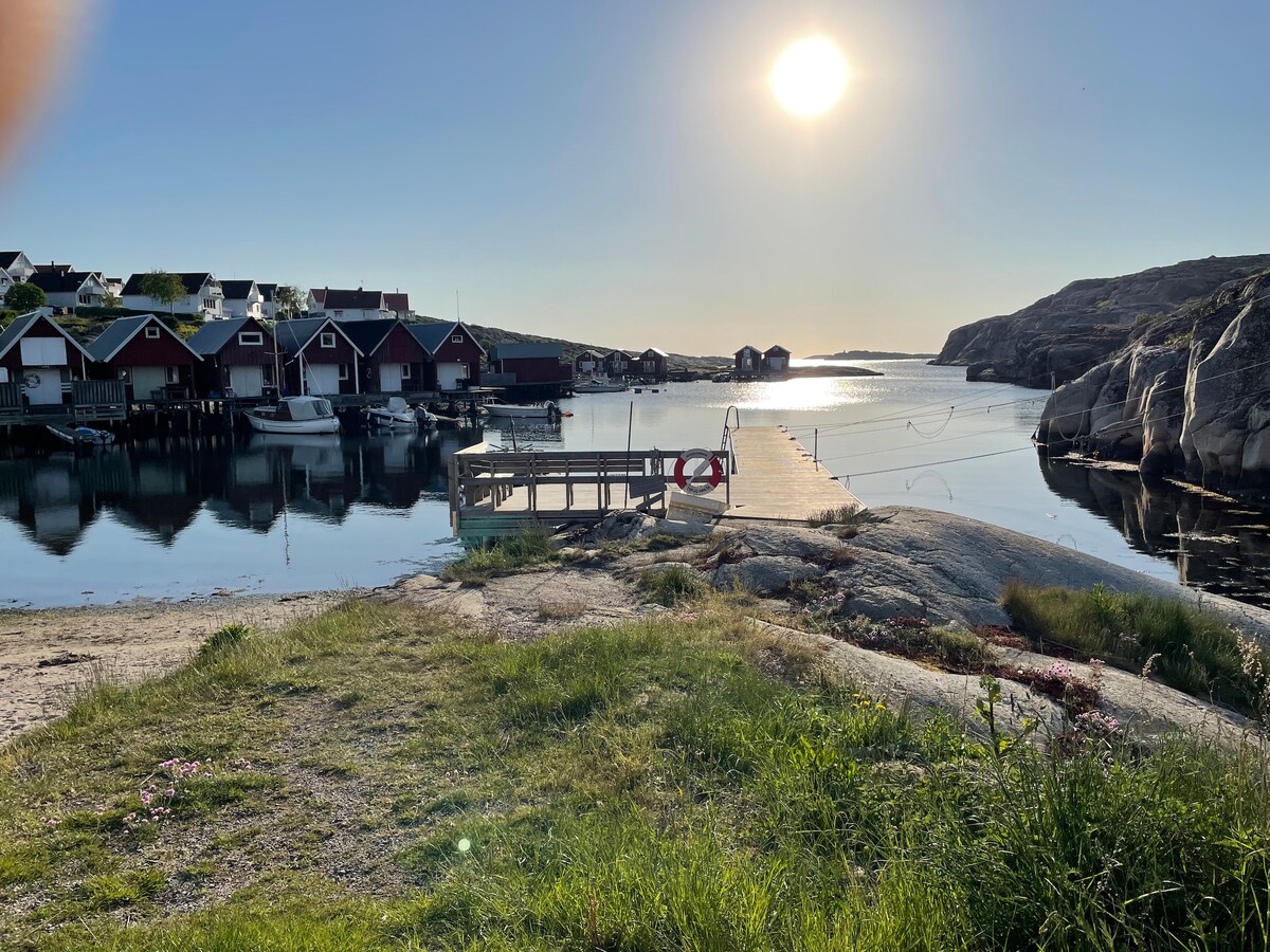 A serene waterfront view is presented, featuring a wooden dock extending into calm waters. Sunlight reflects off the surface, illuminating nearby boathouses and rocky terrain. The scene captures a peaceful atmosphere, inviting moments of relaxation and leisure by the water.