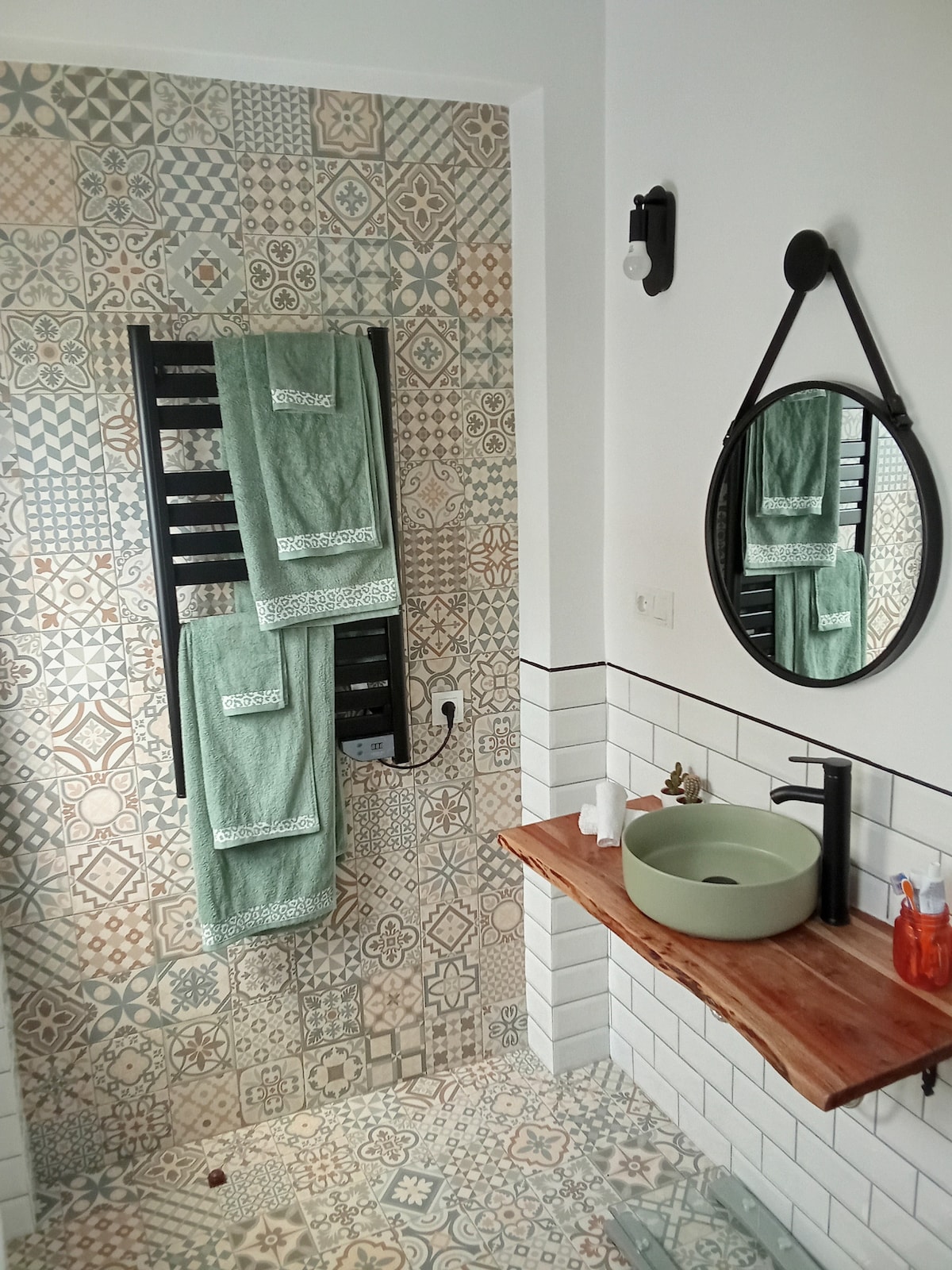 A modern bathroom features intricate patterned tiles in various colors. A round mirror is mounted above a wooden shelf, complemented by a stylish sink. Green towels are hung on a sleek towel radiator, while natural light enhances the space.