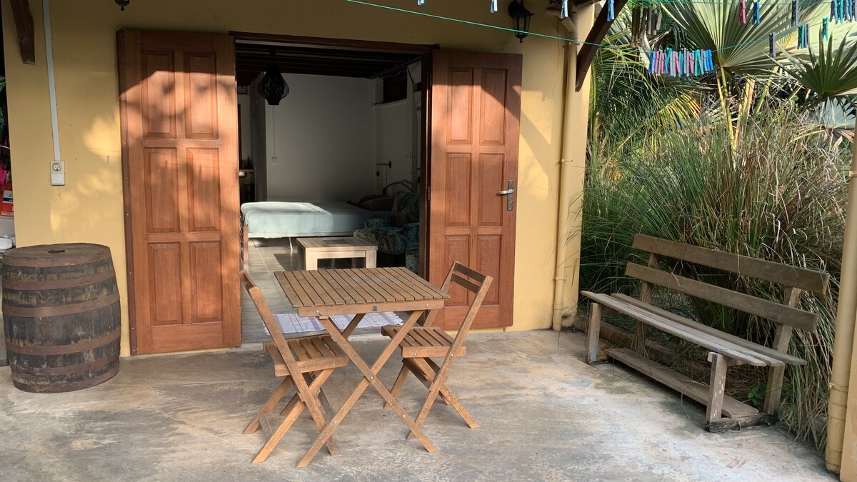 An outdoor space features a wooden table and two folding chairs, inviting relaxation in a tranquil setting. A wooden bench complements the area, while the open doors of the studio reveal a light-colored bed inside. Lush greenery frames the entrance, enhancing the natural ambiance.
