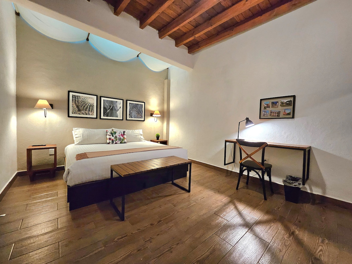 A spacious studio features a comfortable bed dressed with decorative pillows. A wooden bench sits at the foot of the bed. A workspace is provided with a desk and chair, illuminated by a nearby lamp. Wall art and soft lighting create a calm ambiance.