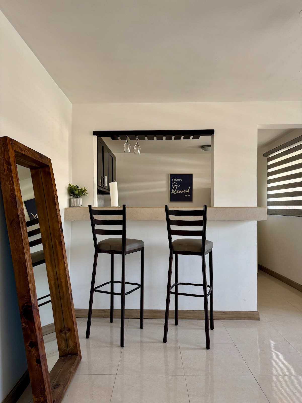 A bright and airy kitchen area features a bar with two high stool seats. A tall mirror stands nearby, reflecting the natural light. A small decorative plant and a candle are placed on the bar. The wall showcases contemporary artwork, enhancing the modern aesthetic.