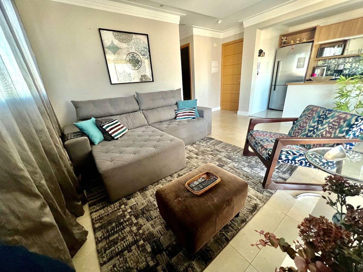 A cozy seating area features a sectional sofa with decorative cushions, a patterned chair, and a small ottoman. Natural light filters through sheer curtains, enhancing the warm tones of the room. A decorative rug adds texture underfoot, while the background includes glimpses of an open kitchen.