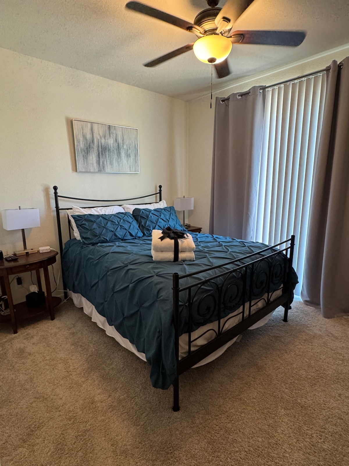A cozy bedroom features a queen-sized bed covered in a teal comforter. Soft, neutral carpet lies beneath, and natural light filters through vertical blinds. Two bedside lamps provide illumination, while a piece of artwork hangs above the bed. A ceiling fan offers additional comfort.