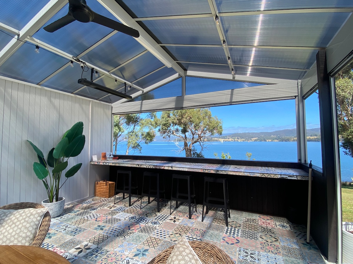 An enclosed alfresco area features a long bar with seating, panoramic views of the water and mountain landscape. The space is decorated with colorful tiles and includes a prominent indoor plant. Natural light floods in through large windows, enhancing the inviting atmosphere.