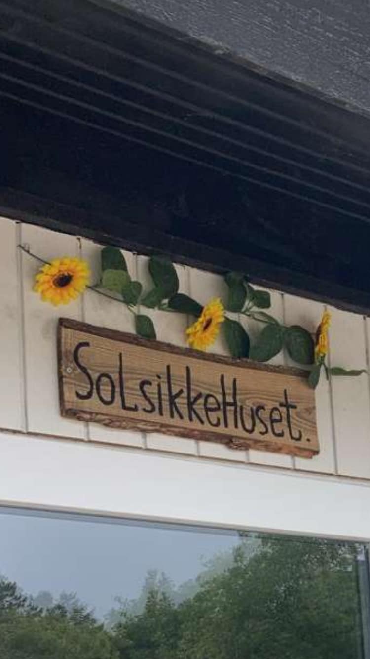 A wooden sign displaying the name 'SolSikkeHuset' is mounted above a glass entrance. Sunflowers are attached to the sign with green leaves, providing a cheerful touch to the rustic decor.