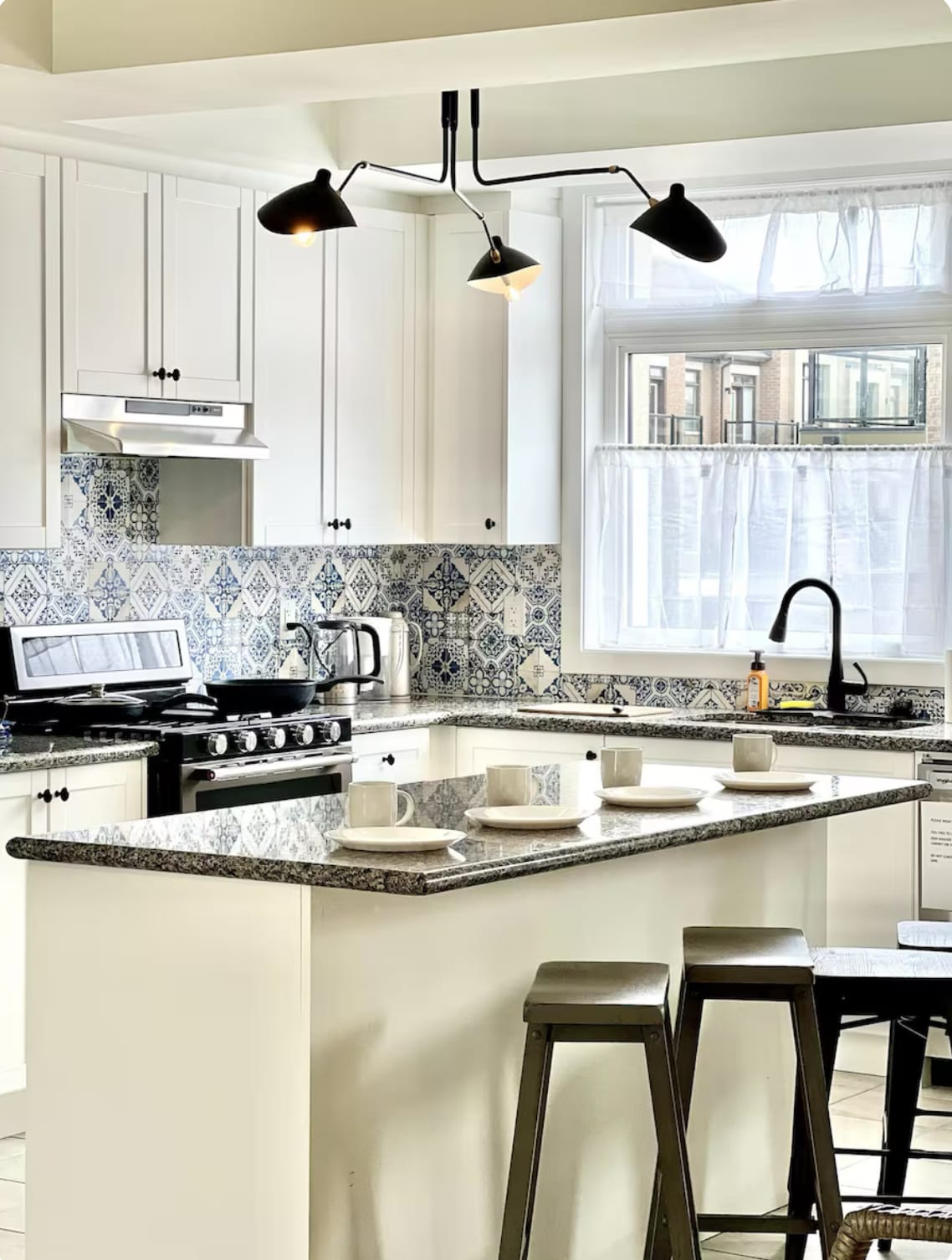 A modern kitchen is presented with white cabinetry and a patterned tile backsplash. A spacious countertop accommodates white dishware arranged neatly. A sleek black faucet and a stylish light fixture enhance the contemporary design. Natural light filters through a window above the sink.