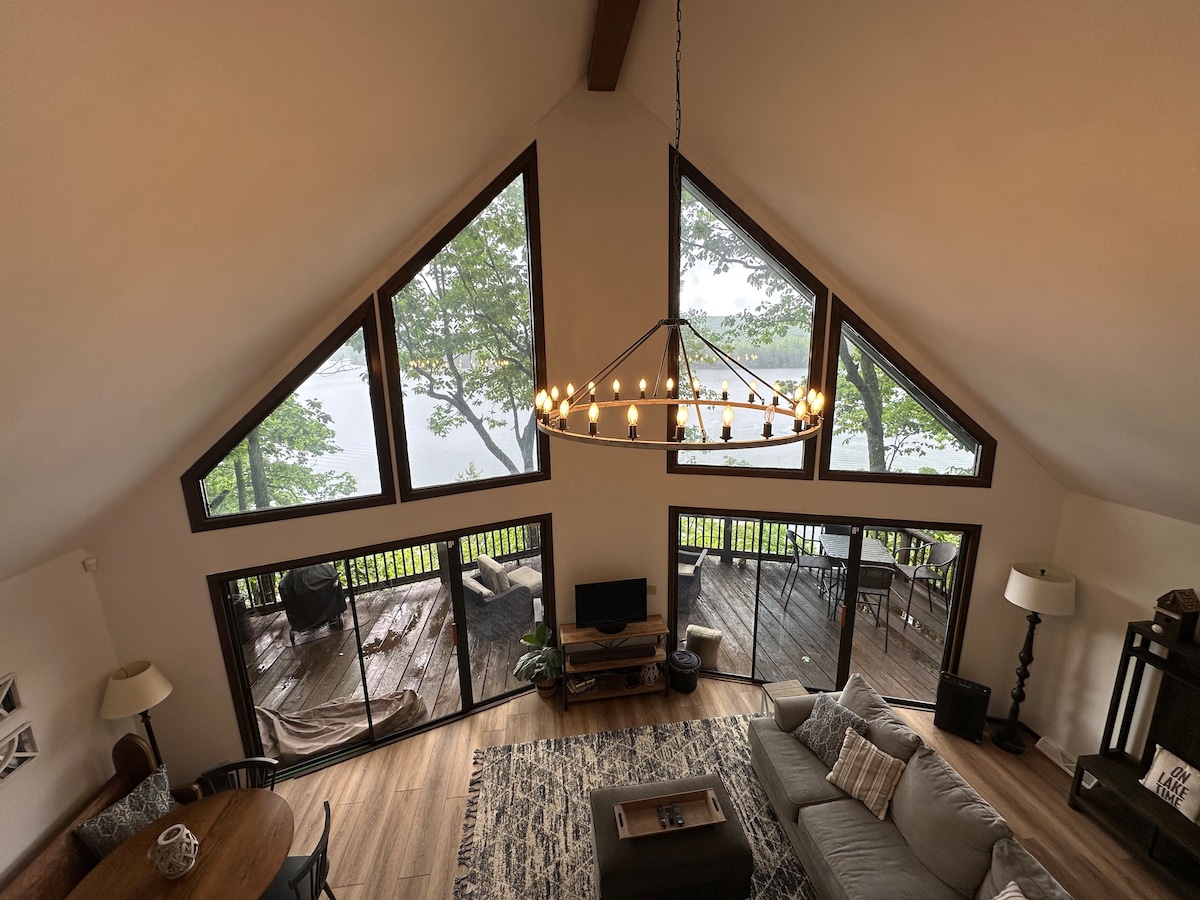 The spacious living area features a high vaulted ceiling, showcasing large triangular windows that frame serene lake views. A cozy sectional couch is positioned in front of a television, while a wooden dining table and an elegant chandelier enhance the inviting atmosphere.