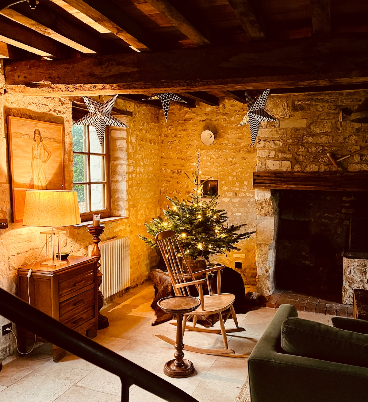 A warm living area features a rustic stone wall and wooden beams, with a small Christmas tree placed next to a rocking chair. Soft light from a lamp illuminates the room, creating a cozy atmosphere. A fireplace is visible, adding to the inviting setting.