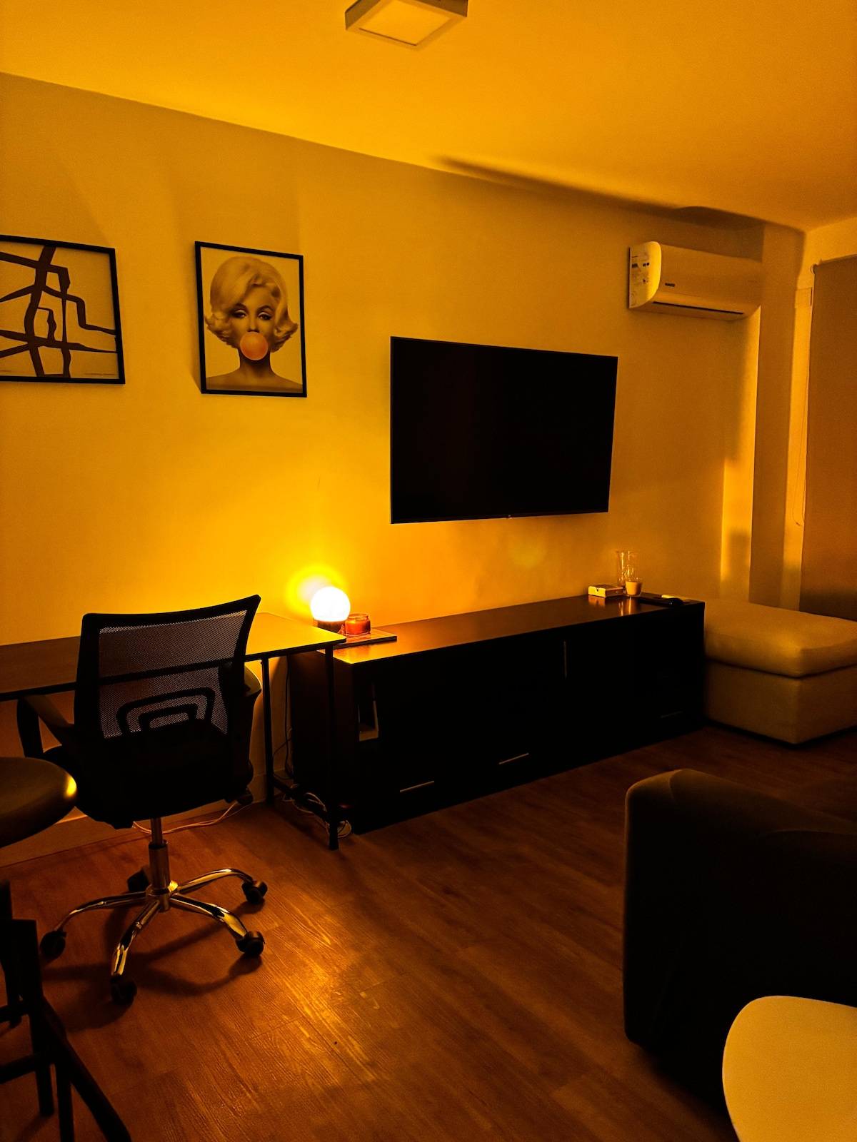 A cozy living area is illuminated by soft lighting. A mounted flat-screen television is positioned above a sleek dark surface, alongside a stylish lamp. A comfortable chair and a desk area are present, with hardwood flooring adding warmth to the space.