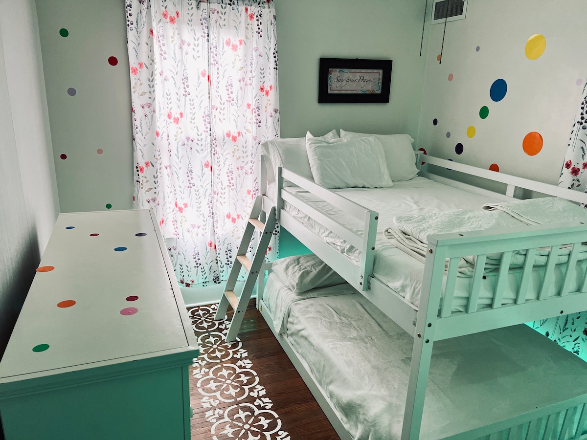 A cheerful children's bedroom is presented with a bunk bed featuring a spacious upper sleeping area and a lower sleeping area. Colorful polka dots adorn the walls, complemented by bright, patterned curtains letting in natural light. A white dresser sits against the wall, enhancing the playful ambiance.