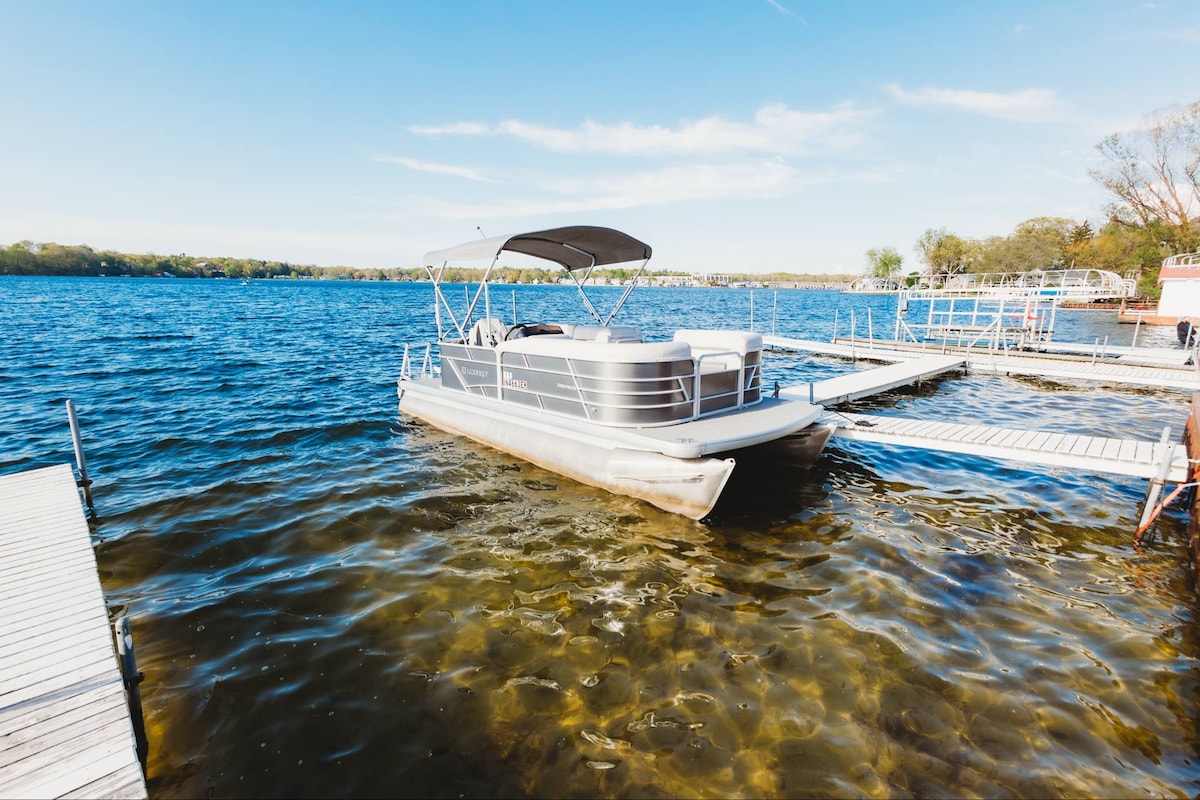 A pontoon boat is docked in calm waters, surrounded by wooden docks. The clear water exhibits a gentle gradient, showcasing sandy bottom beneath. The horizon is visible under a clear blue sky, emphasizing a serene waterfront setting.