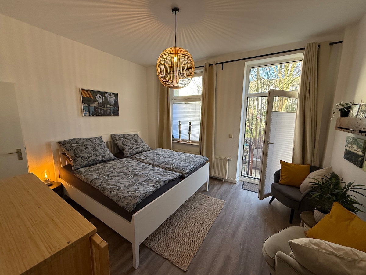 The spacious bedroom features two beds with patterned bedding, accompanied by a cozy armchair and a small side table. Natural light floods the room through a large window and glass doors leading to a terrace, enhancing the connection to the outdoor space.
