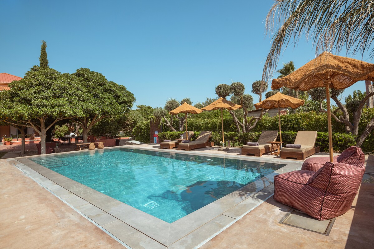 A refreshing swimming pool is showcased amidst lush greenery, with sun loungers and umbrellas positioned around the edges. The pool area is designed for relaxation, providing space for guests to unwind in a serene outdoor setting.