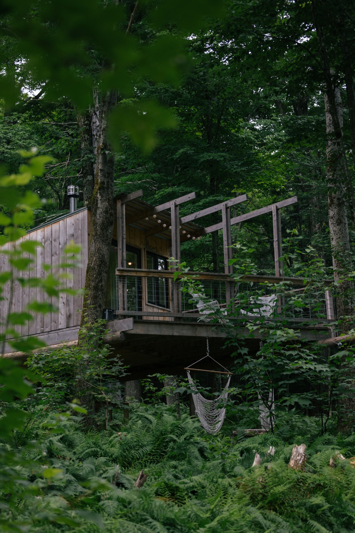 The Treehut is nestled among lush greenery, featuring a wooden exterior that integrates with its natural surroundings. The structure is elevated and supported by sturdy beams, with a deck showcasing seating options. A hanging macrame piece adds a decorative touch beneath the structure.