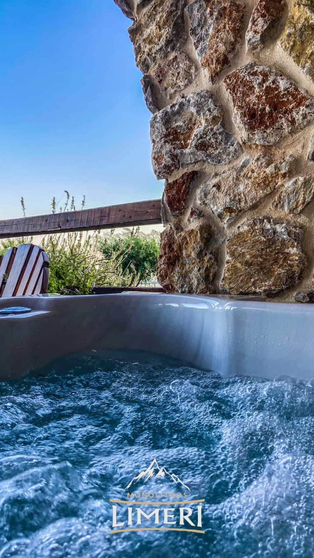 A relaxing outdoor jacuzzi is framed by a rustic stone wall. The bubbling water is highlighted against a clear blue sky, with greenery visible in the background, creating a serene setting for unwinding.