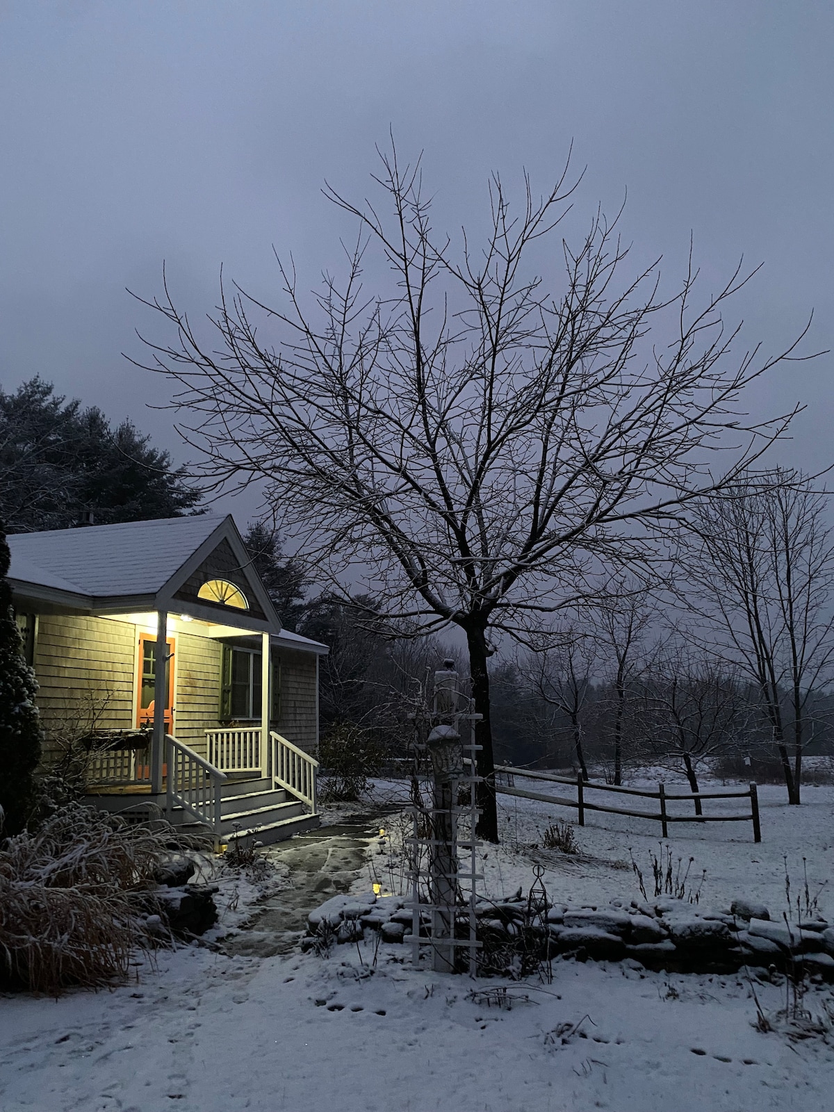 A charming cottage is presented under a soft blanket of snow in the evening. Warm light spills from the entrance, illuminating the porch and pathway. Leafless trees and a fence define the serene landscape, creating a peaceful winter ambiance.