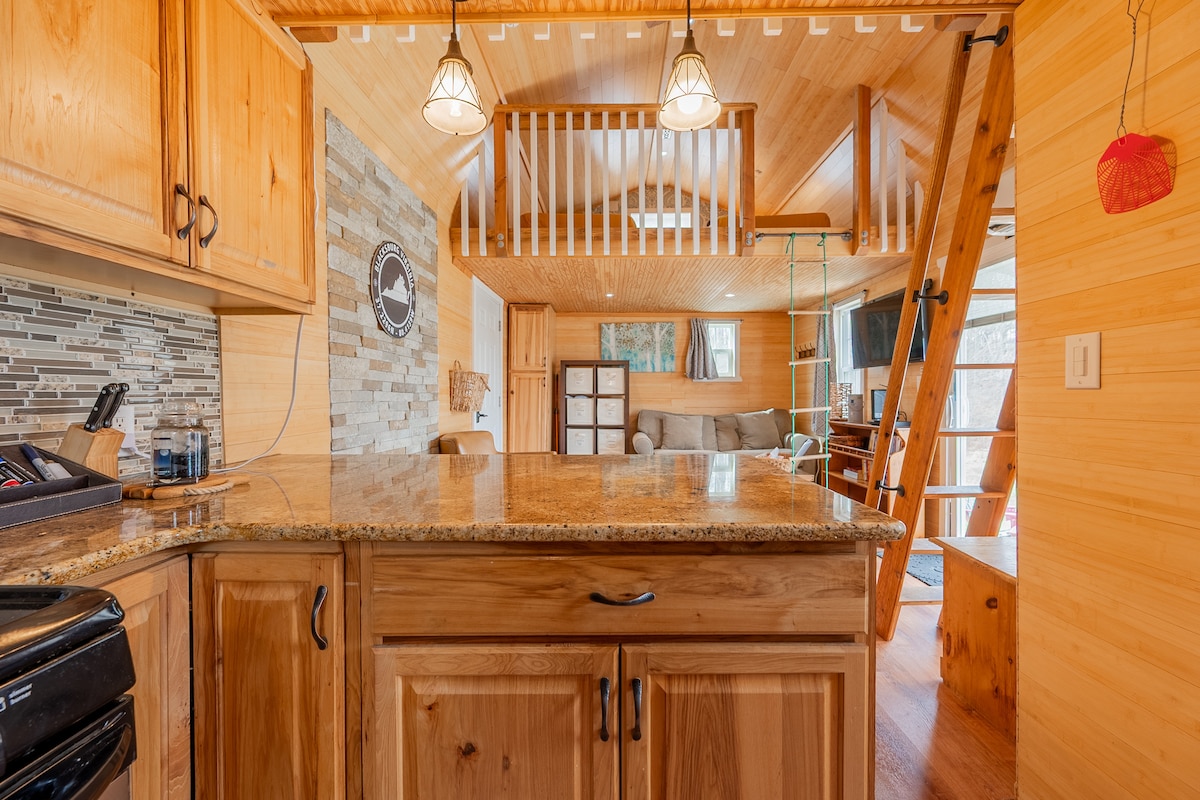 The modern kitchen features hickory cabinetry and a granite countertop with bar seating for two. A tiled backsplash complements the space, while a rustic accent wall adds warmth. A wooden ladder and loft area are visible in the background, overlooking the living space.