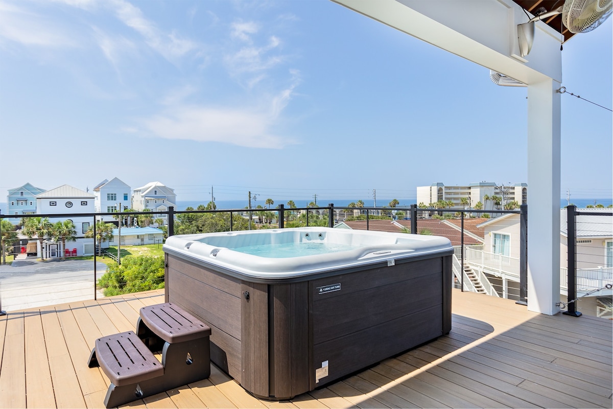 A spacious rooftop deck features a six-person hot tub surrounded by a wooden deck. The sky is clear, providing unobstructed views of the Gulf and nearby coastal homes, creating an inviting space for relaxation.