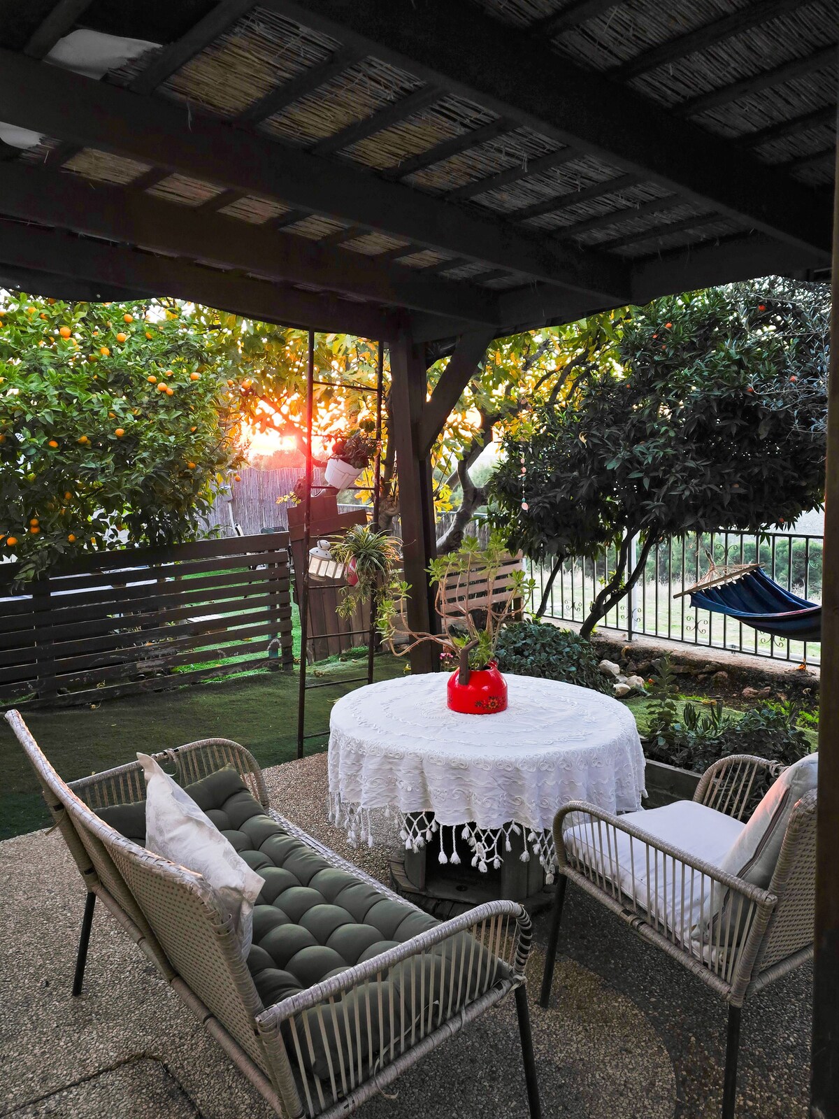An outdoor seating area features a round table with a white tablecloth, surrounded by two chairs. Lush greenery is present, offering a tranquil atmosphere, while the sun sets in the background, illuminating the space with a warm glow.