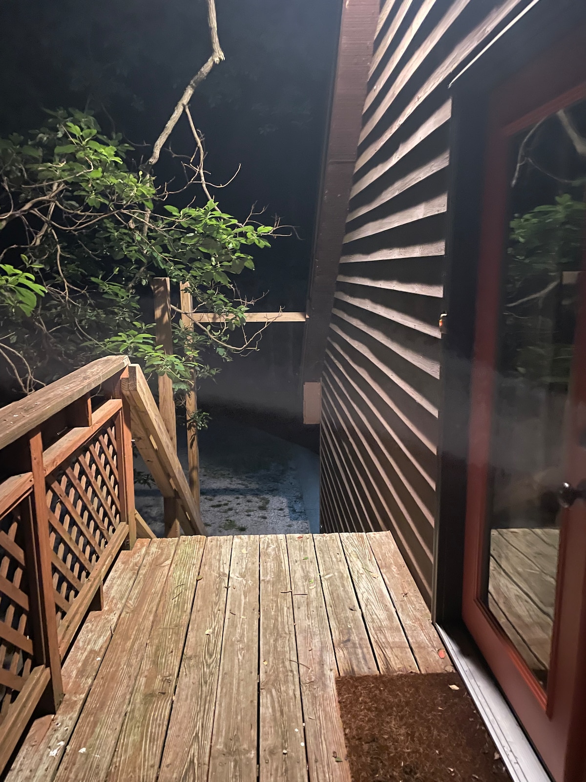 The exterior entrance features a wooden deck leading to a staircase surrounded by natural greenery. Soft lighting highlights the textured wooden siding of the building, while a glimpse of a wooded area is visible in the background.