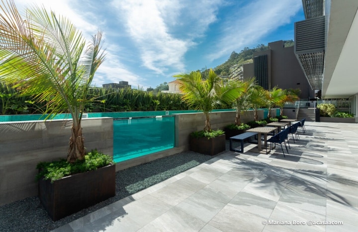 A serene outdoor area is featured, showcasing a modern pool lined with tall tropical plants. Comfortable seating is available on a stone patio, with surfaces reflecting natural light. The backdrop includes greenery and architectural elements, enhancing the tranquil ambiance.