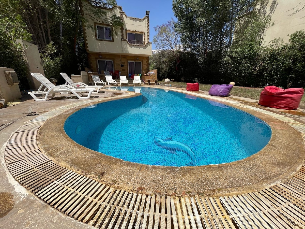 A private pool, shaped uniquely, is surrounded by a paved patio. Several sun loungers are positioned around the pool, complemented by colorful bean bag chairs placed in the grassy area nearby. Lush greenery provides a natural backdrop to the tranquil setting.