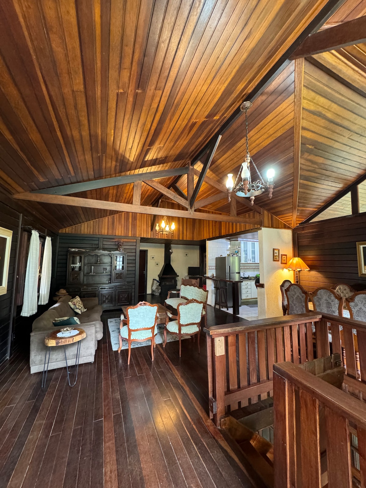 A cozy living area is characterized by rich wooden walls and a high ceiling. Comfortable seating is arranged around a central coffee table, while a rustic fireplace offers warmth. Light fixtures and ambient decor enhance the inviting atmosphere of the space.