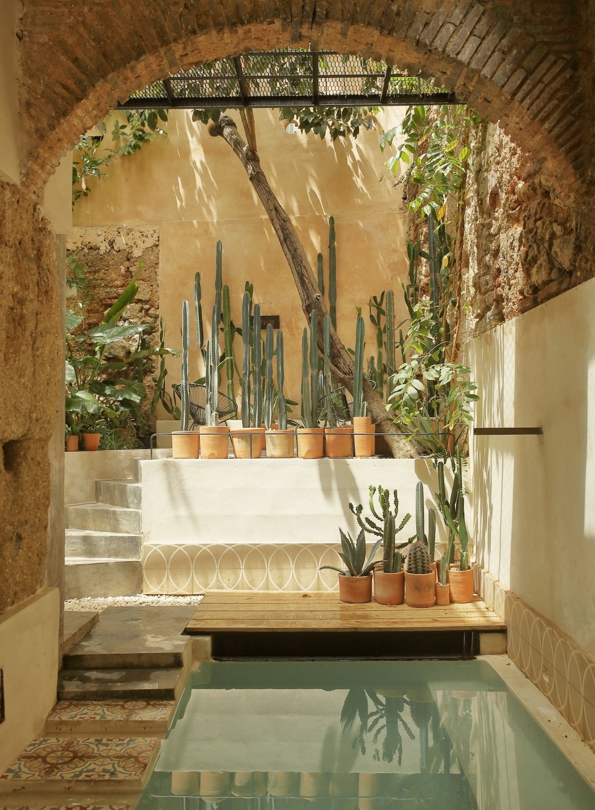 A serene courtyard is framed by an original archway, featuring a shallow reflection pool surrounded by potted cacti. Sunlight filters down, highlighting vibrant greenery against the rustic walls, contributing to a tranquil and inviting atmosphere.